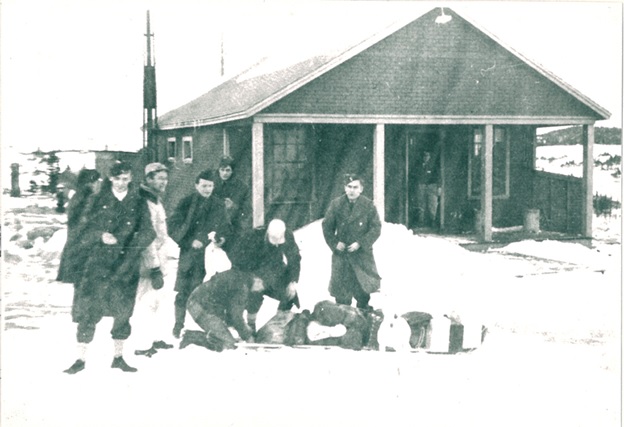 Small group of airmen prepared to walk across the “Barrens” home for Christmas leave. Foreground Marvin Miller behind the group, Norman Paquet at the right, behind the toboggan buttoning his coat. Man with the white cap is Ed Flowers watching Mickey Stevens attaching the luggage to the toboggan.